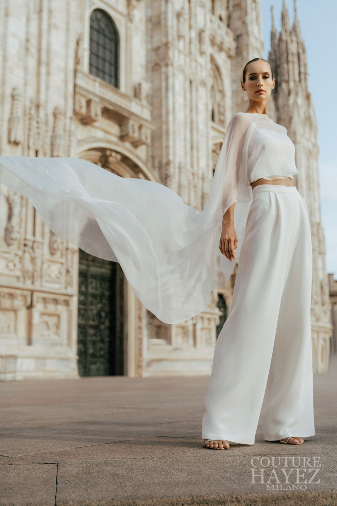 Modella con mantella lunga, top in pizzo e pantaloni palazzo Couture Hayez – Atelier sposa Milano