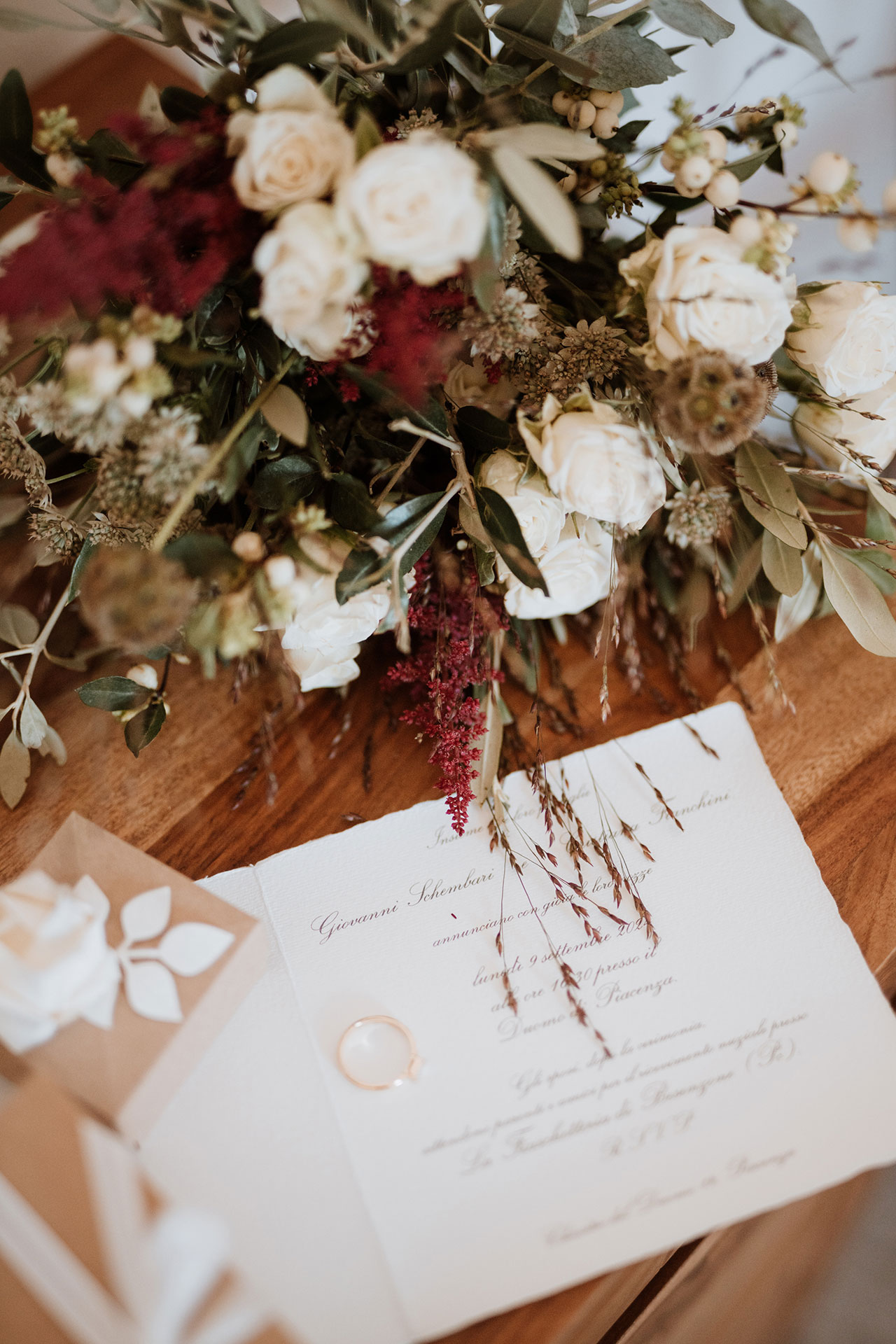 Bouquet da sposa con rose bianche, eucalipto, amaranto bordeaux, fedi nuziali e partecipazione di matrimonio calligrafica
