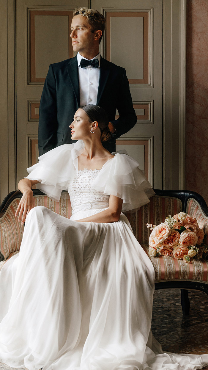 sposa-e-sposo-in-villa-sul-lago-di-como sposa con abito morbido in seta seduta su divanetto di una villa del lago di Como con sposo accanto in piedi